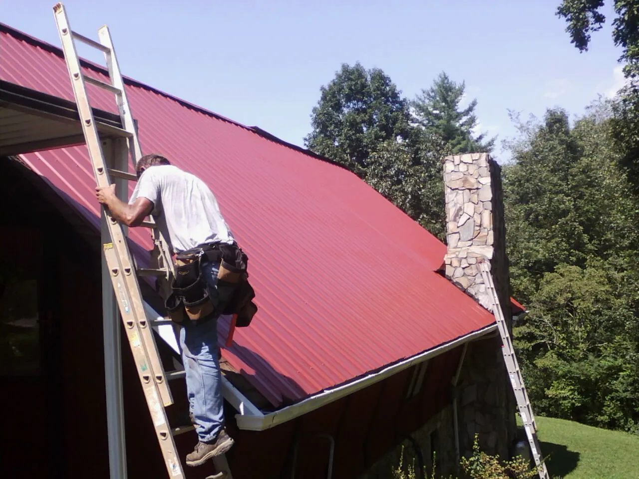Roofer on a ladder beside a red metal roof and stone chimney — Soffit & Fascia Repair by Roofing Brothers in Preston Hollow